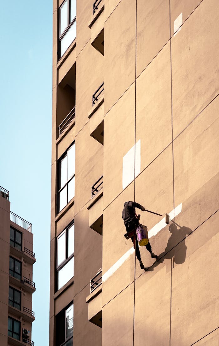 Services Man painting tall building exterior while suspended outdoors, showcasing urban maintenance work.
